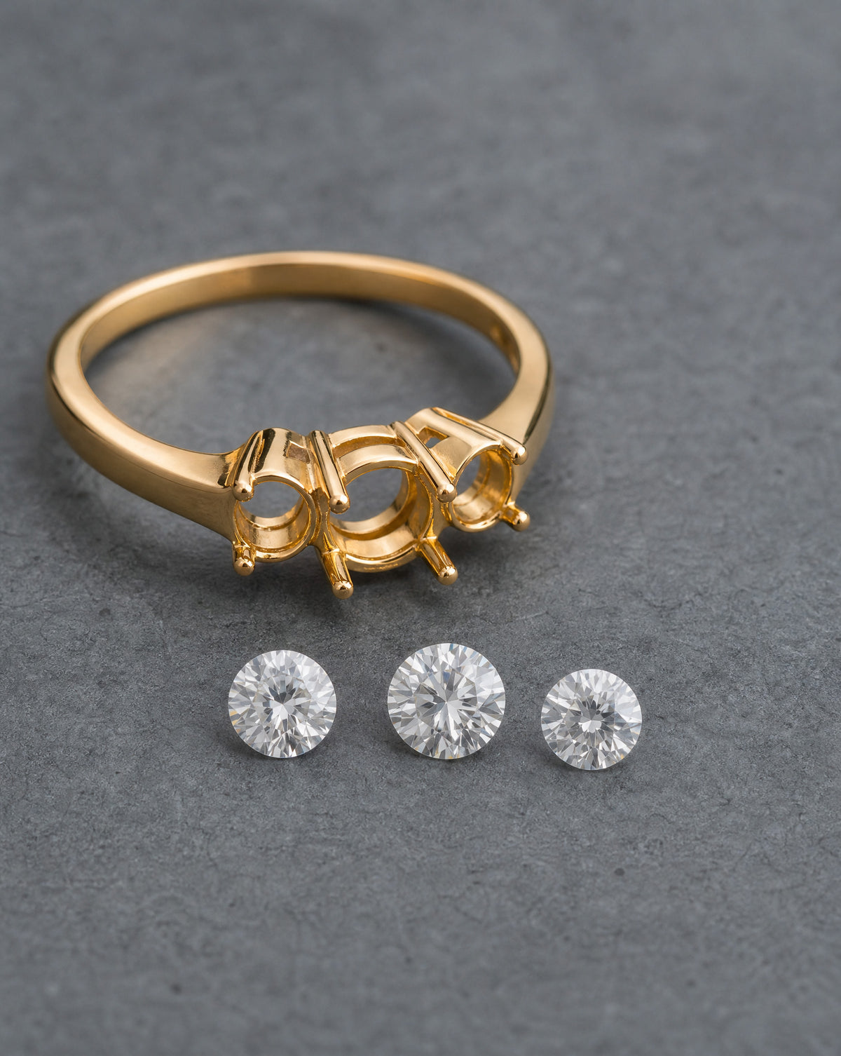 A gold ring setting with three prongs and three round clear gemstones displayed below it on a textured gray background.