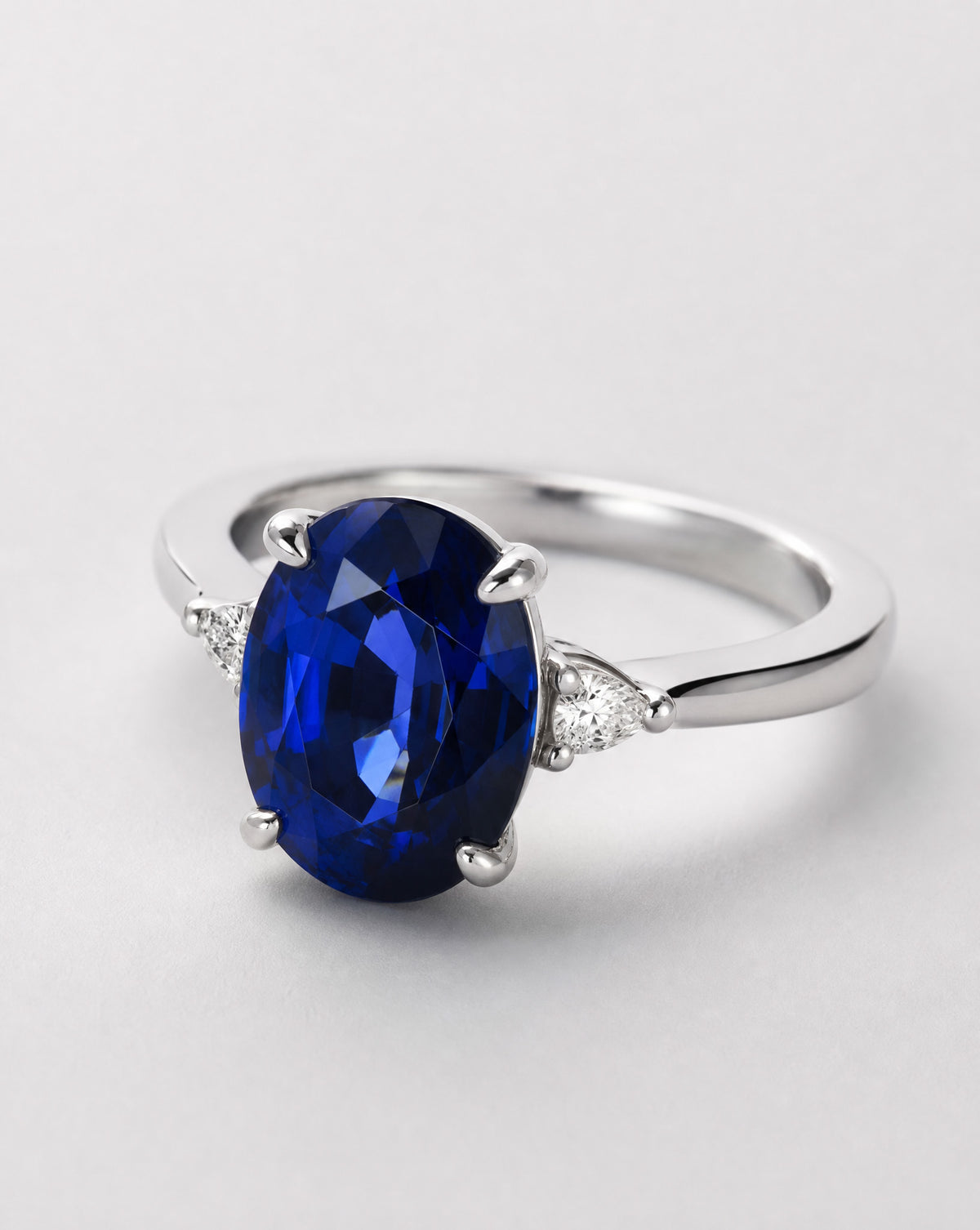 An oval-cut deep blue sapphire ring set in a polished silver-colored band with small triangular clear stones on either side, photographed on a light gray background.m