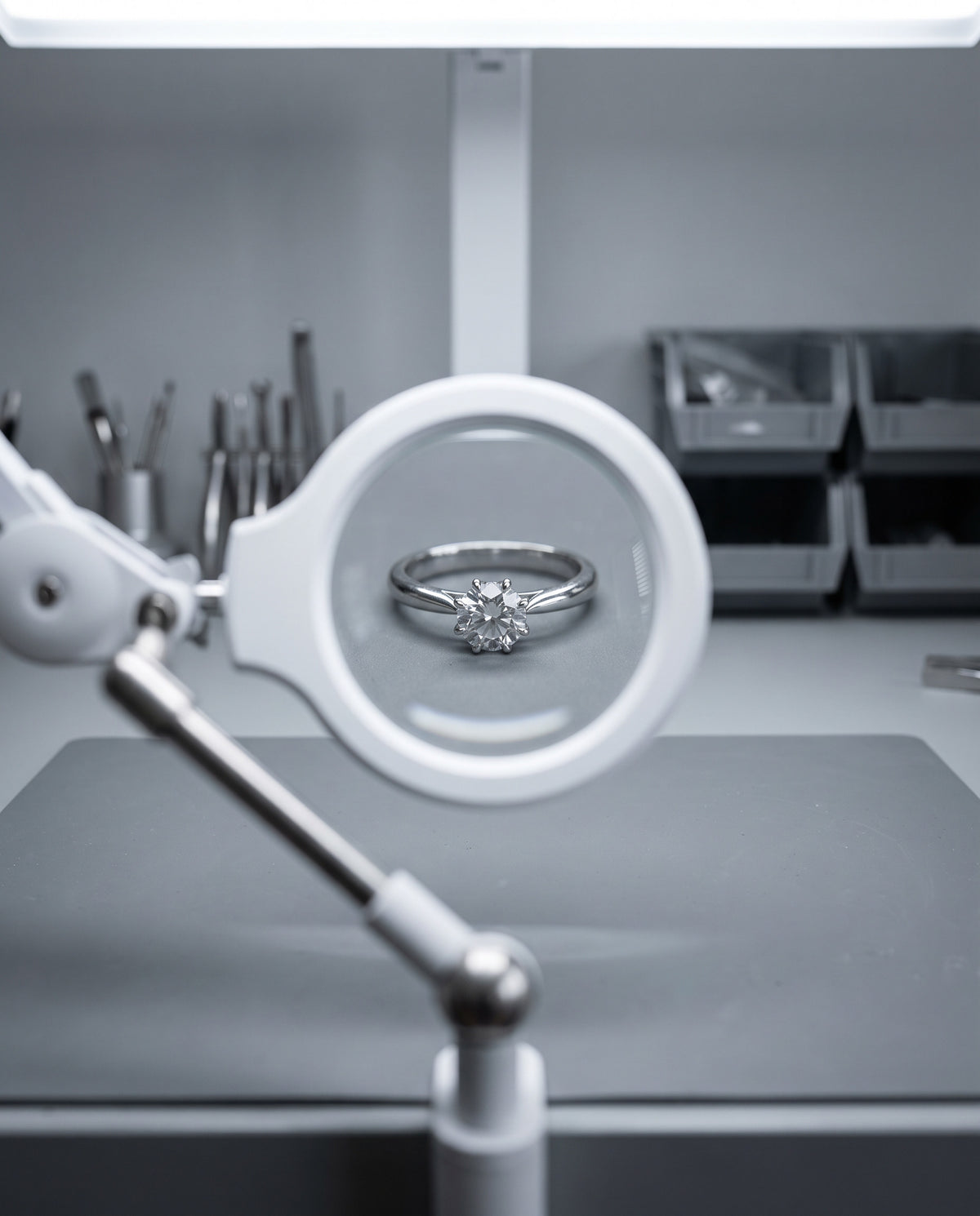 A silver ring with a floral cluster centerstone is viewed through a white magnifying loupe mounted on an adjustable stand, set against a gray workshop background with tools and storage bins.