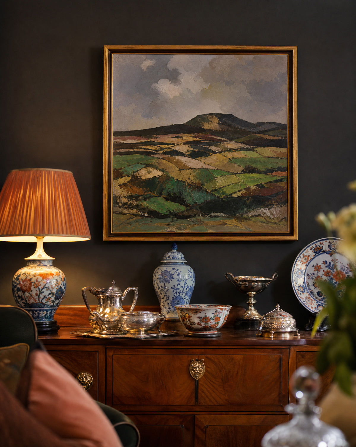 A framed landscape painting of rolling green and brown hills under a cloudy sky, displayed on a dark wall above a wooden console table with a lamp, teapot, and decorative dishes.