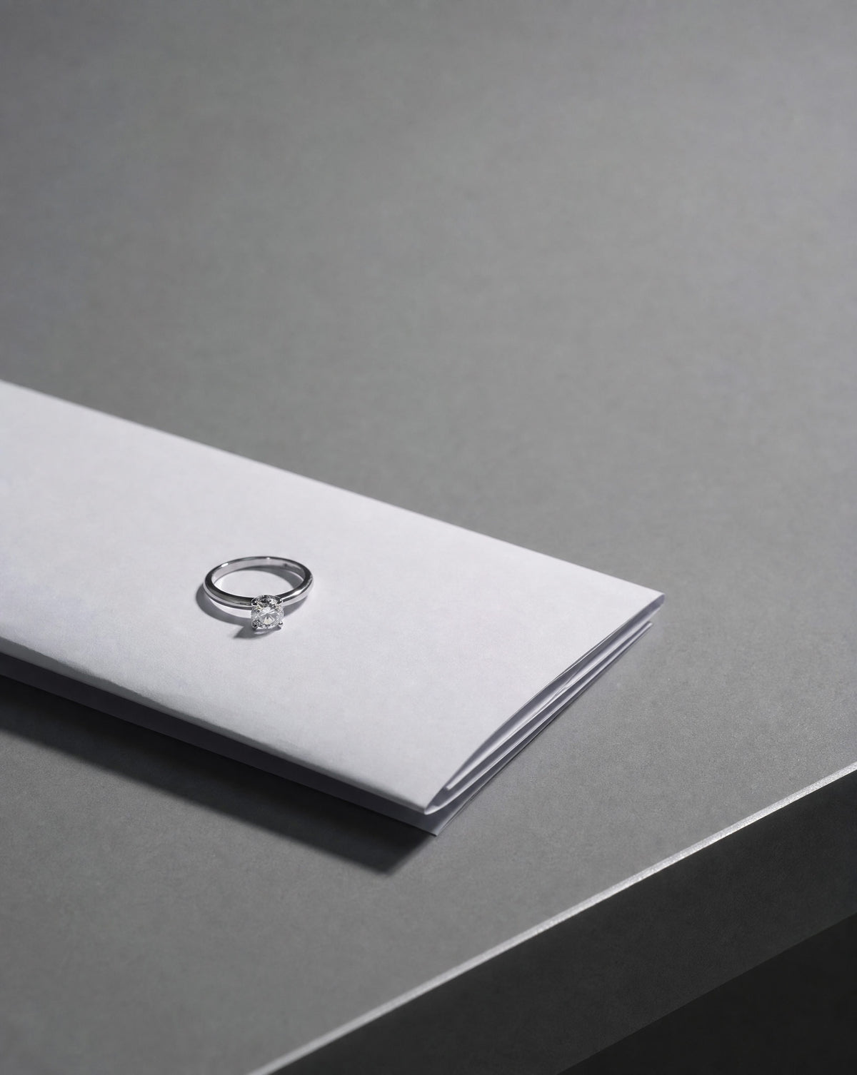 A silver ring with a small clear gemstone, displayed on top of a folded white paper envelope on a dark gray surface.