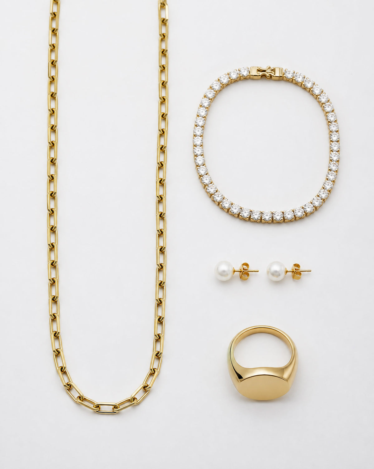 A gold jewelry set on a white background, including a long curb chain necklace, a tennis bracelet with clear stones, a signet ring with a white pearl, and a pair of pearl stud earrings with gold settings.