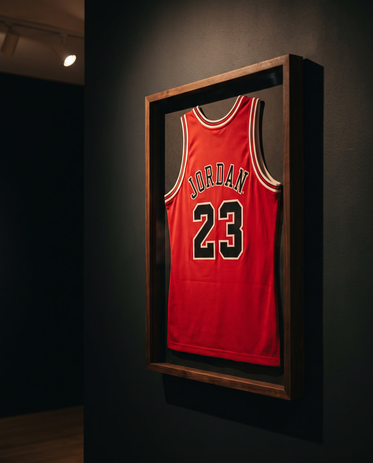A framed red Chicago Bulls Michael Jordan number 23 basketball jersey hangs on a dark wall in a gallery or showroom.
