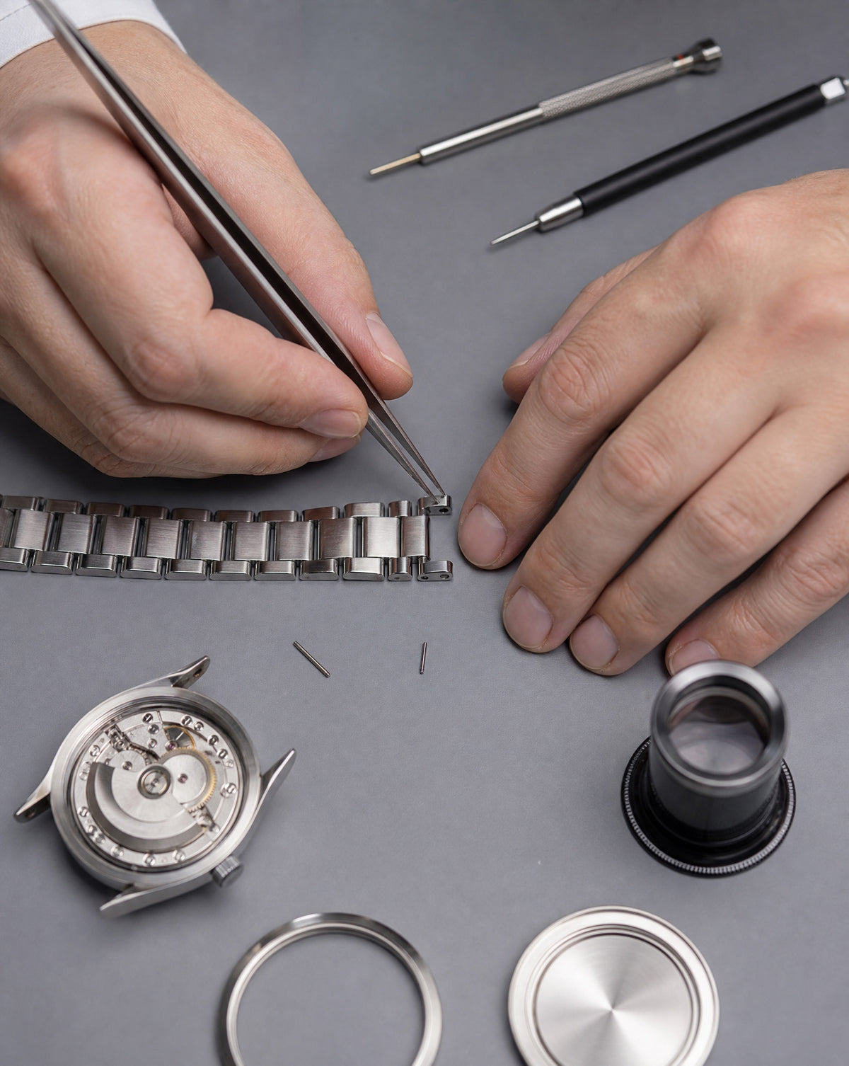Close-up of a watchmaker's hands using tweezers to place a silver metal watch band next to a disassembled silver watch case, with tools and rings arranged on a gray surface.